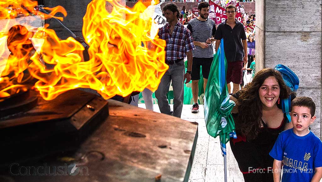 Con una agitada manifestación de trasfondo, la madre se detiene unos minutos para explicar a su hijo de qué se trata la llama votiva. Luego continuará con la protesta.